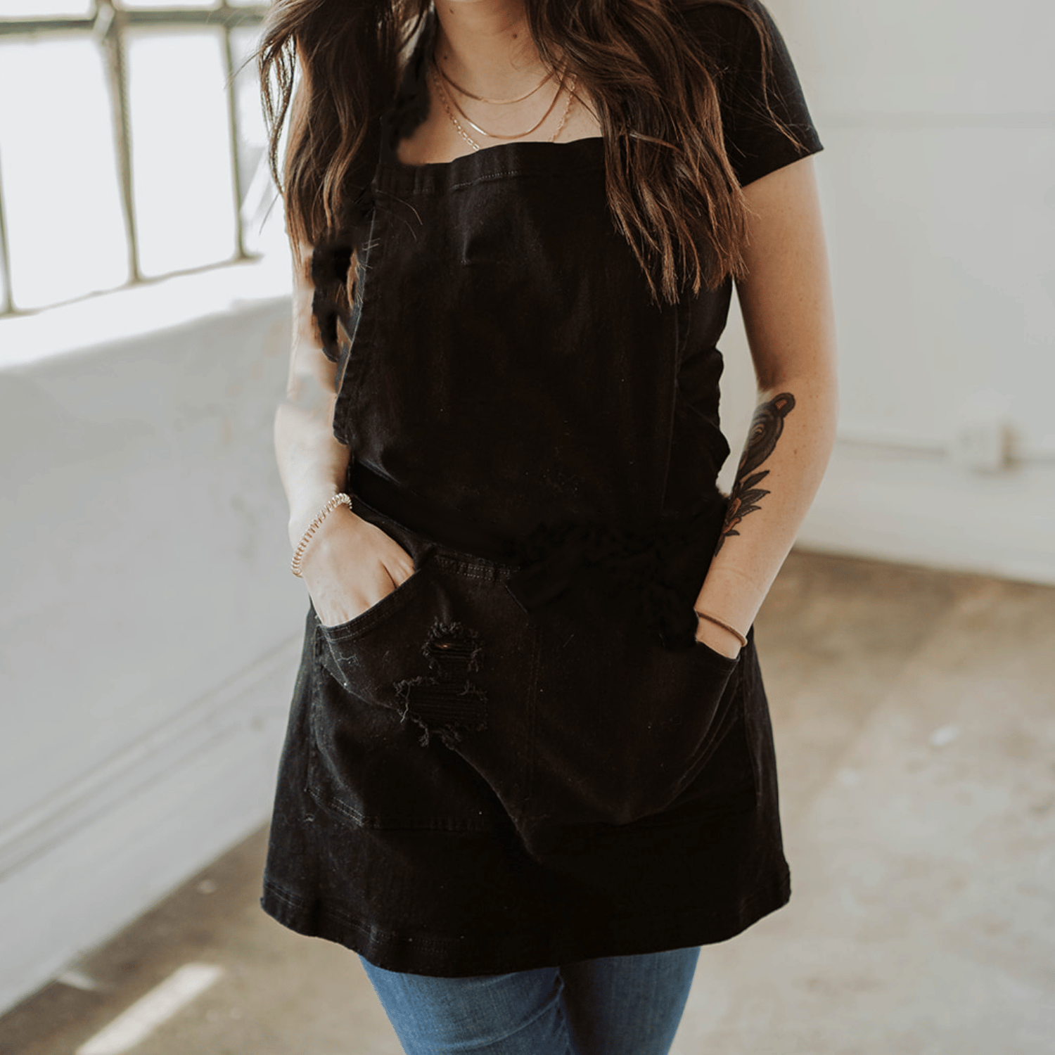 Pure black distressed denim salon apron by J Clark Designed displayed on female model with her hands inside apron pockets.