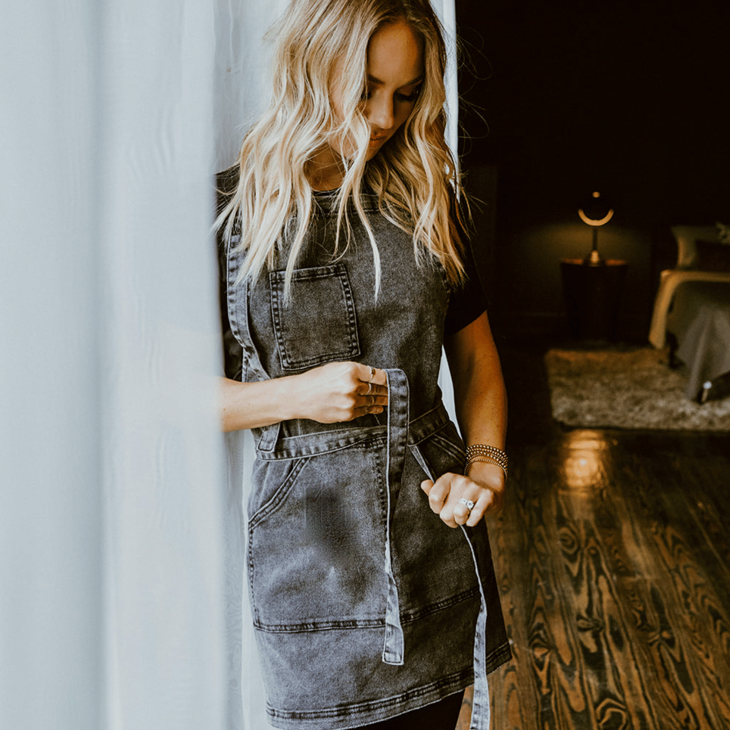Woman wearing the Over Shoulder Black Acid Washed Apron by J Clark Designed while adjusting the waist ties.