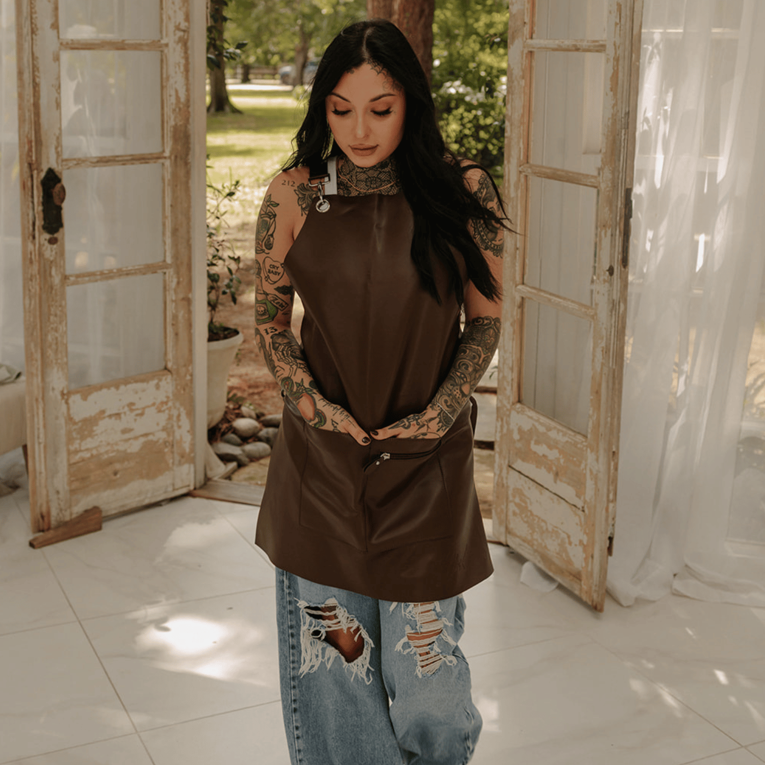 Female model walking while looking at the floor wearing ripped wide leg jeans and the Cocoa Brown Leather apron by J Clark Designed. Her hands are in the two front waist pockets.