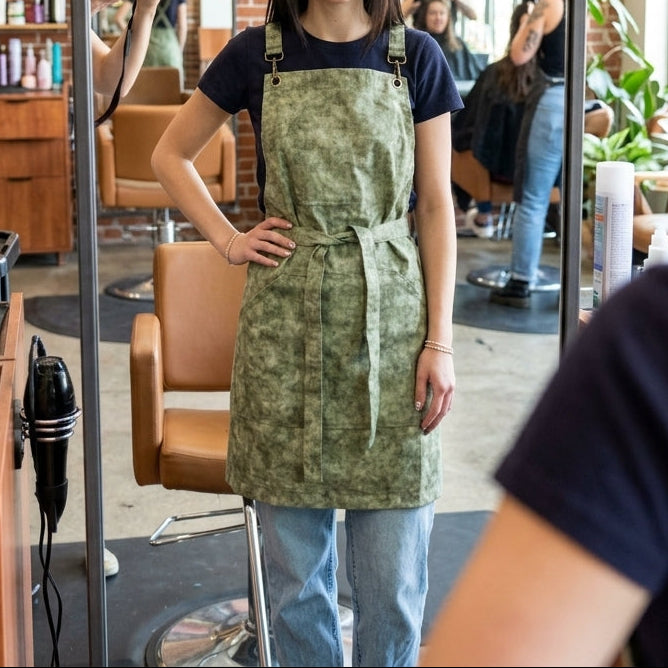 Woman in a green apron standing in a hair salon with mirrors and hair products.