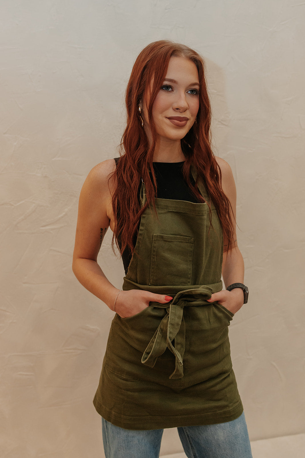 Olive green apron by J Clark Designed worn by petite hair stylist with her hands in the apron pockets.