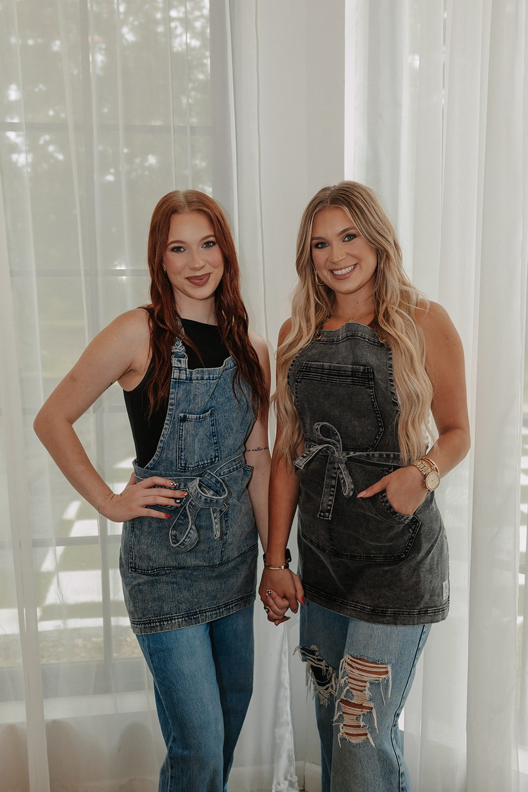 Two female models wearing J Clark Designed Aprons. Model on the left is wearing the Denim Distressed Apron. Model on the right is wearing the Over Shoulder Black Acid Washed Apron.