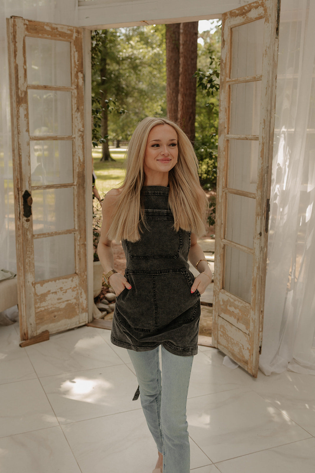 Female model wearing the Over Shoulder Black Acid Washed apron by J Clark Designed as she walks through a doorway with her hands in the apron pockets.