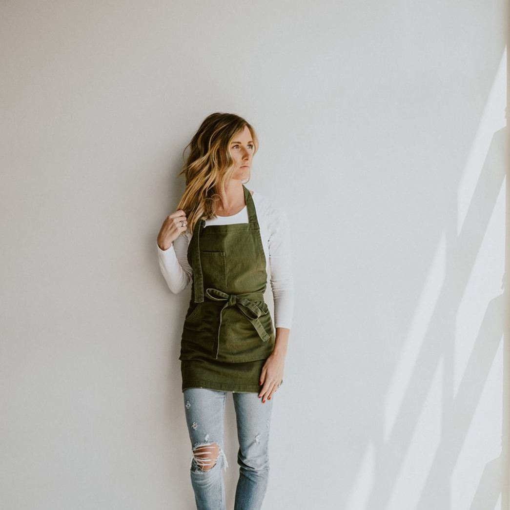 Olive Green apron by J Clark designed worn by female model leaning back against a white wall.