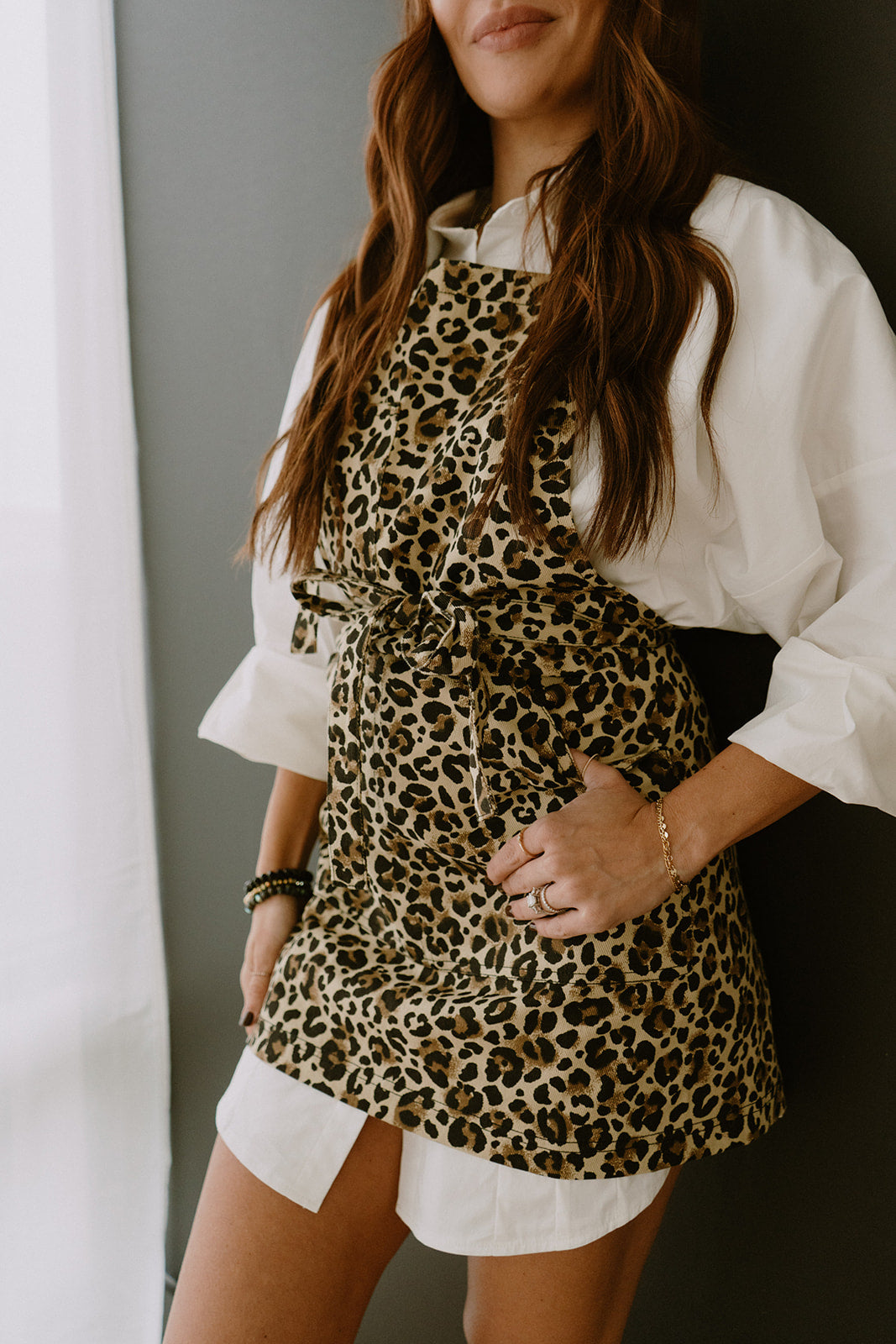 Female model leaning against a gray wall wearing an oversized white dress shirt and the Leo apron by J Clark Designed.