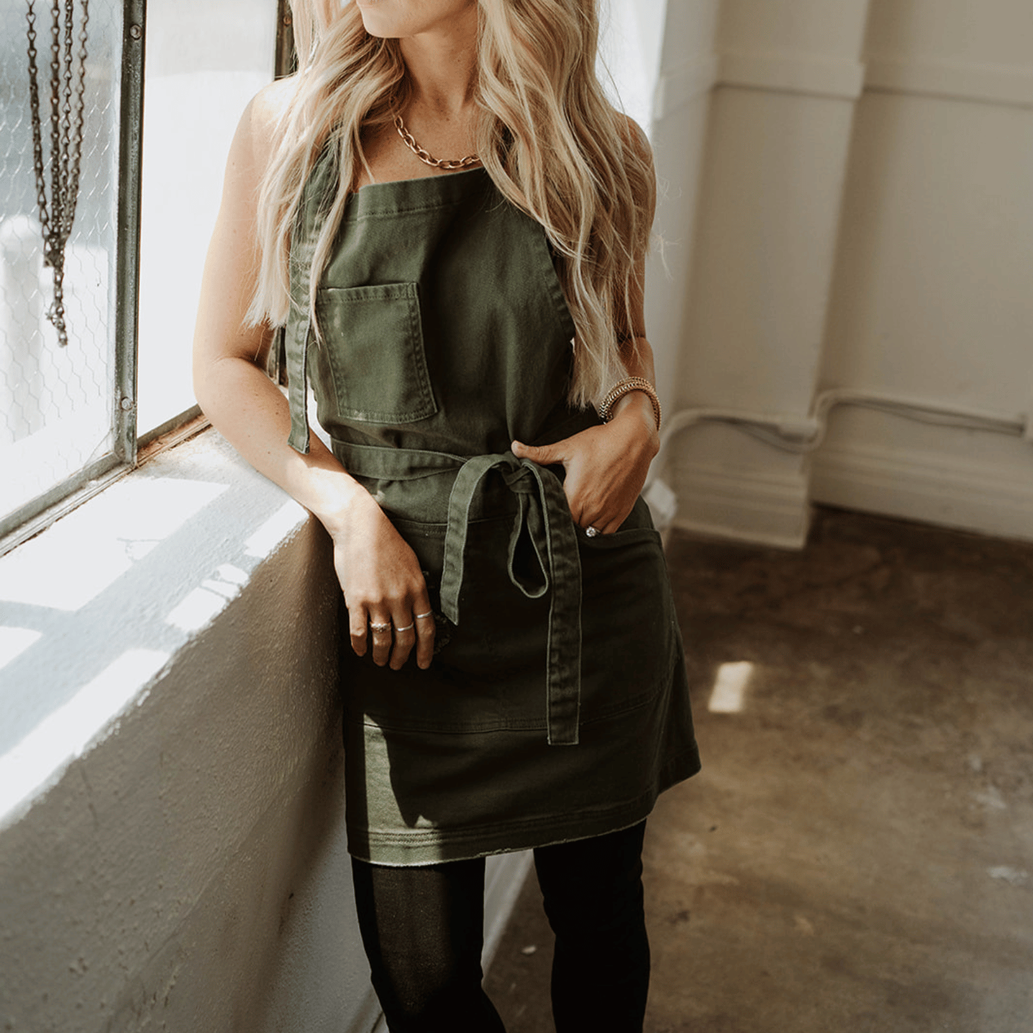 Female model leaning on window sill wearing the Olive Green Apron by J Clark Designed.