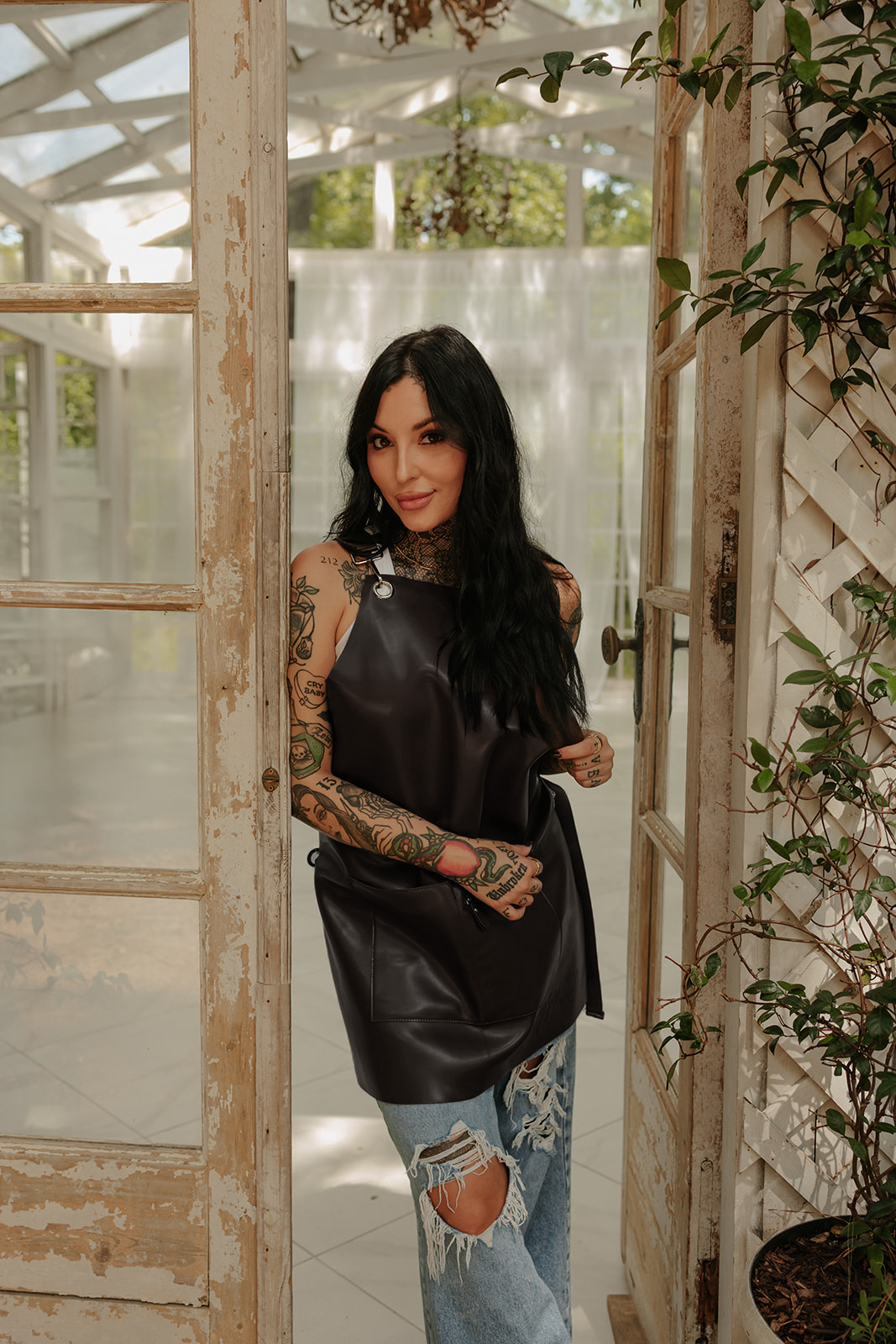 Full black vegan leather bleach proof salon apron By Jclark Designed worn by a young woman standing in a doorway.
